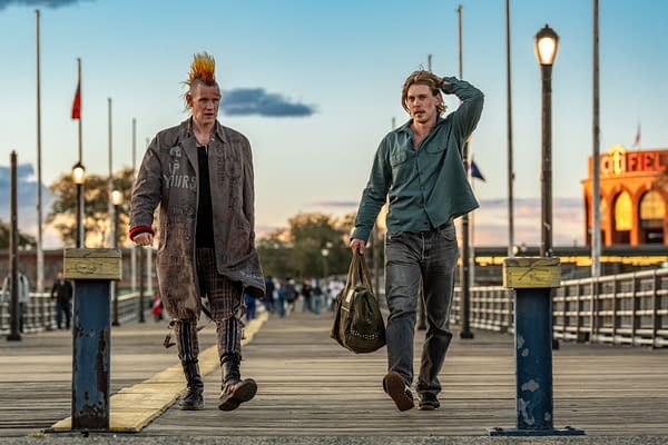 Two young men walk along a wooden pier during sunset, one with bright orange spiked hair and a heavily graffitied oversized coat, while the other wears a green shirt and carries a bag. The scene captures a casual, urban atmosphere.