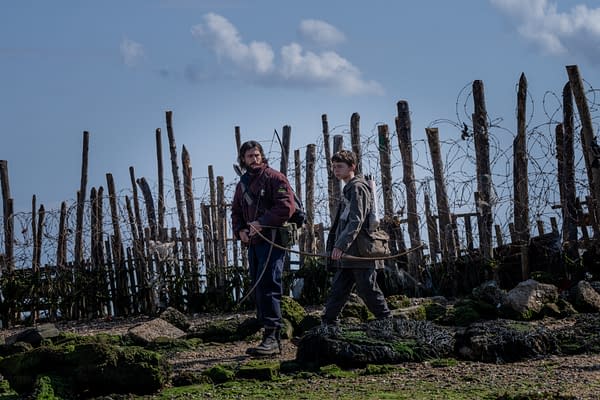 Two characters walk along a rocky landscape near a barbed wire fence, with a sparse backdrop of clouds in a clear sky. The figures appear equipped for survival, suggesting a post-apocalyptic setting.