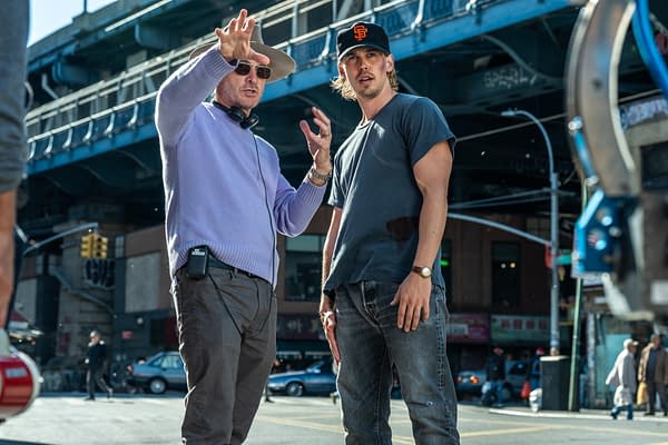 Behind-the-scenes moment featuring a director in a lavender sweater and sunglasses gesturing while discussing scenes with a male actor in a gray t-shirt and baseball cap, set against an urban backdrop with streets and buildings.