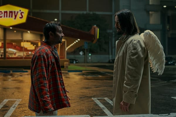 A scene from the film "Good Fortune" featuring Aziz Ansari as Arj and Keanu Reeves as Gabriel, who appears with angel wings. The setting is a dimly lit parking lot with a Denny's restaurant in the background.