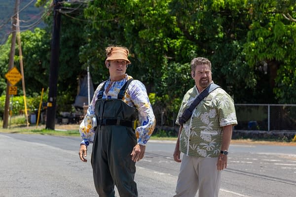 Live-action scene from Disney's 'Lilo & Stitch' featuring Billy Magnussen as Pleakley, wearing a colorful floral shirt and overalls, alongside Zach Galifianakis as Jumba, dressed in a green Hawaiian shirt, standing on a roadside with lush greenery in the background.