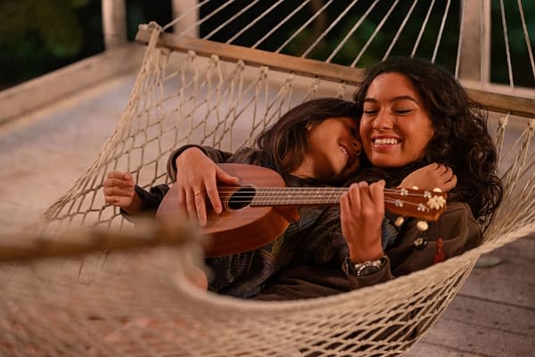 Two young women relax together in a hammock, with one playing a ukulele. The scene captures a warm and intimate moment, highlighting the bond between Lilo and her older sister Nani from Disney's live-action adaptation of 'Lilo & Stitch.'