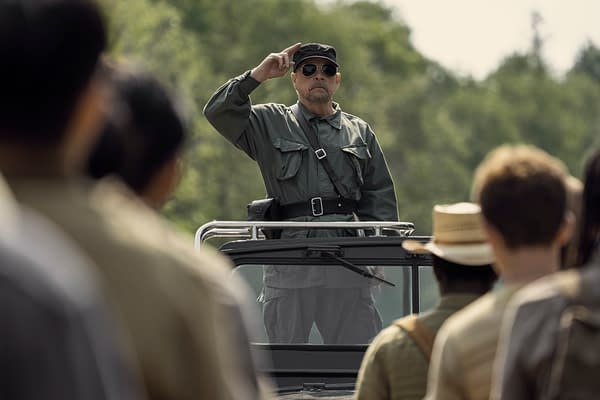Mark Hamill portrays The Major in a scene from 'The Long Walk,' standing on a vehicle and saluting a group of individuals in military attire, with a wooded background.