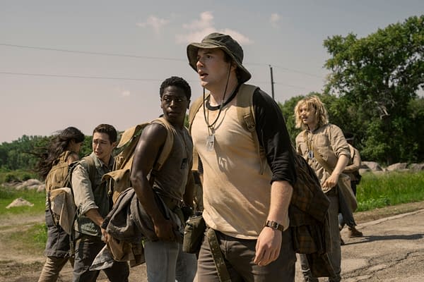 A scene from 'The Long Walk' featuring a group of young characters, including a diverse cast, walking along a rural path with trees and rocks in the background. They appear determined and focused on their journey.