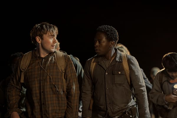 Cooper Hoffman as Garraty and David Jonsson as McVries appear to have an intense conversation while walking in a dark setting in 'The Long Walk.' They are wearing backpacks and surrounded by other figures in the background.