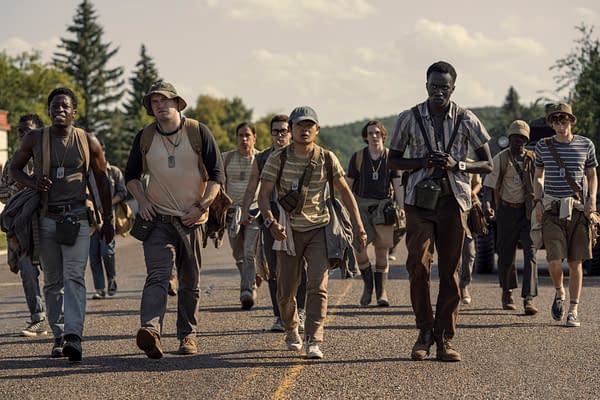 A group of young men, including characters portrayed by David Jonsson, Cooper Hoffman, and Jordan Gonzalez, are walking together along a road, dressed in casual outdoor clothing with backpacks and gear. The scene reflects a tense atmosphere as they embark on a significant journey in The Long Walk.
