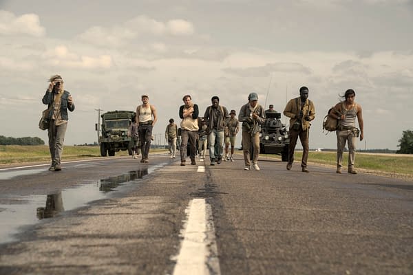 A scene from 'The Long Walk' featuring a group of young men walking on a highway, with military vehicles in the background. The characters appear determined as they traverse the road under a cloudy sky.