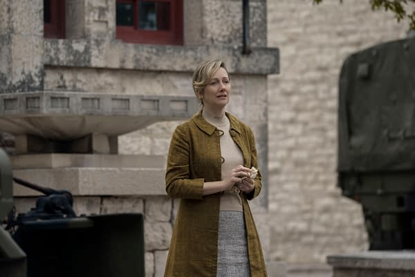 Judy Greer as Ginny Garraty in The Long Walk, standing outside near a stone wall, wearing a mustard-colored coat and holding something in her hands.