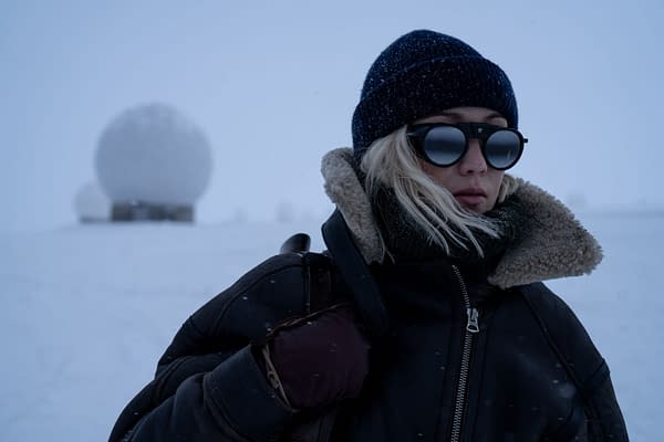 In a snowy landscape, Pom Klementieff portrays the character Paris from 'Mission: Impossible - The Final Reckoning,' wearing dark sunglasses and a warm, lined jacket, with a knitted beanie, against a backdrop of spherical snow-covered structures.
