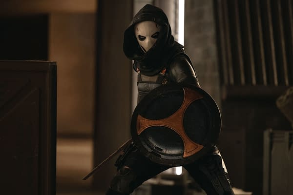 A character dressed as Taskmaster, portrayed by Olga Kurylenko, wears a hood and a mask while holding a shield, in a scene from Marvel Studios' THUNDERBOLTS*. The background features industrial elements.