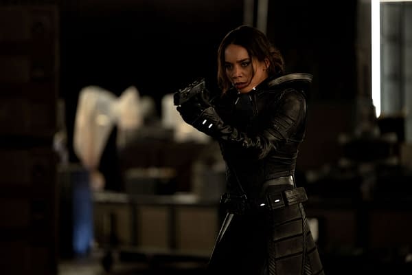 A character portraying Ghost, played by Hannah John-Kamen, stands ready with a firearm, dressed in a sleek black suit with intricate detailing. The background suggests an industrial setting with shadows and muted lighting, typical of Marvel Studios' atmosphere.
