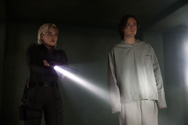 In a scene from Marvel Studios' THUNDERBOLTS*, Yelena Belova, portrayed by Florence Pugh, stands with a flashlight aimed while Bob, played by Lewis Pullman, appears apprehensive beside her. They are positioned in a dimly lit setting that adds to the tension of the moment.