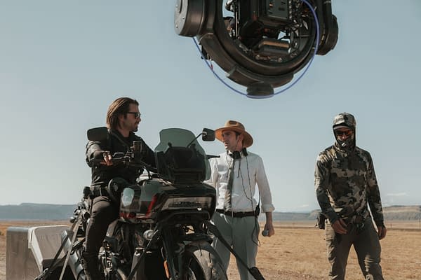 On the set of Marvel Studios' 'Thunderbolts,' actor Sebastian Stan sits on a motorcycle while director Jake Schreier, wearing a hat and holding a microphone, discusses the scene with a crew member in camouflage attire. The setting features an open landscape under a clear sky.