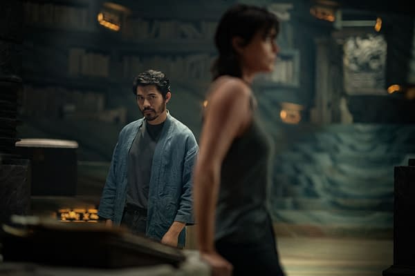 A scene from 'The Old Guard 2' featuring Henry Golding as Tuah in a blue robe, looking intently at Charlize Theron as Andy, who is in the foreground. The setting is dimly lit with shelves of books in the background.