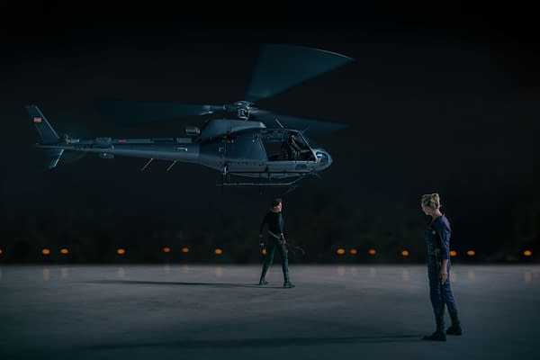A dramatic scene from 'The Old Guard 2' featuring a helicopter hovering above two characters, one standing with a weapon, and the other looking down. The atmosphere is tense and dark, highlighting the intense action of the film.