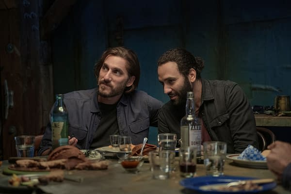 A scene from The Old Guard 2 featuring Luca Marinelli as Nicky and Marwan Kenzari as Joe at a dinner table, surrounded by various foods and drinks in a dimly lit setting.