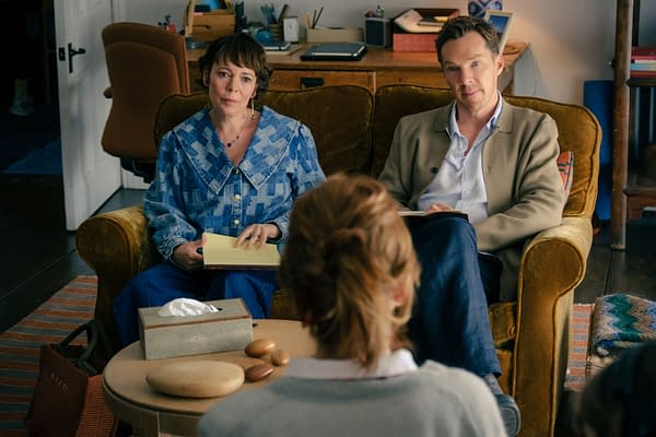 A scene from 'The Roses' featuring Olivia Colman and Benedict Cumberbatch sitting on a couch, engaged in conversation with a person out of frame. The setting includes a cozy living room with a coffee table displaying a tissue box and smooth stones.