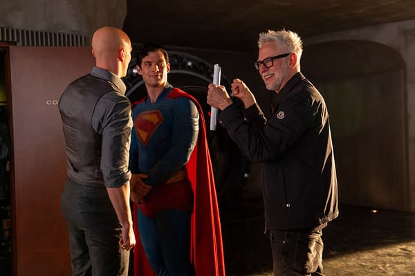 A behind-the-scenes moment featuring two men conversing, one dressed as Superman in a blue suit with a red cape, and a third man smiling and holding a script, likely a director on set.