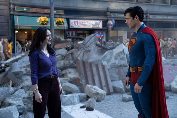 A scene featuring a woman in a purple sweater and a superhero in a blue suit with a red cape, standing amidst rubble in an urban setting. The background includes a coffee shop and hints of a crowd, suggesting a post-disaster scenario.