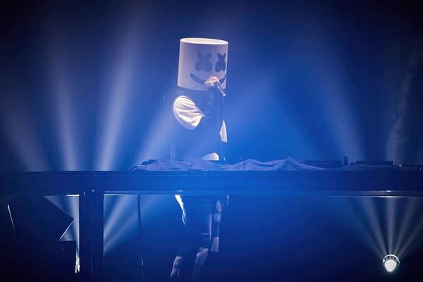 A DJ wearing a distinctive marshmallow-shaped helmet performs on stage, surrounded by dramatic blue lighting during a lively event. The scene captures the festive atmosphere of a concert celebrating the holidays.