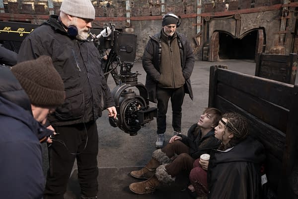 A behind-the-scenes moment from a film set featuring a director in a black jacket with headphones, discussing with two young actors seated on the ground. A large camera setup is visible, along with other crew members in warm clothing.