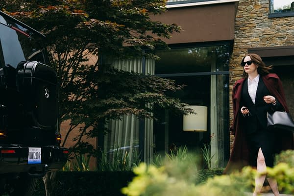 In a scene from 'BUGONIA,' Emma Stone portrays Michelle Fuller, confidently walking outside a modern building dressed in a stylish black suit and a long burgundy coat, with sunglasses and a handbag.
