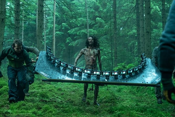 A scene from a film shoot in a dense forest, featuring a partially nude actor covered in dirt and blood, being carried on a large, curved filming rig by crew members. The atmosphere is misty and green, enhancing the eerie setting.