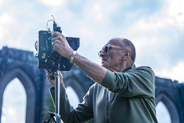 A photographer, wearing sunglasses, is adjusting a camera on a tripod in front of a historic stone archway under a bright sky.