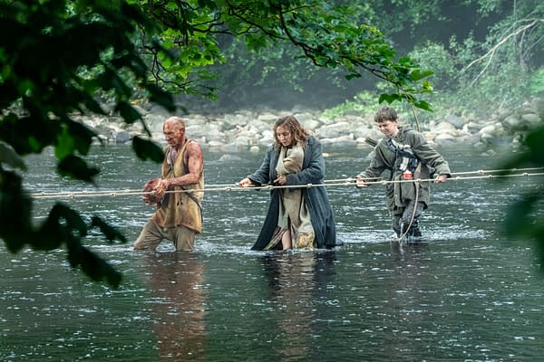 Three characters wade through shallow water, using a rope for support. The scene conveys a sense of survival and struggle, with one individual appearing dirty and rugged, another in a wool coat, and a younger person holding a makeshift weapon.