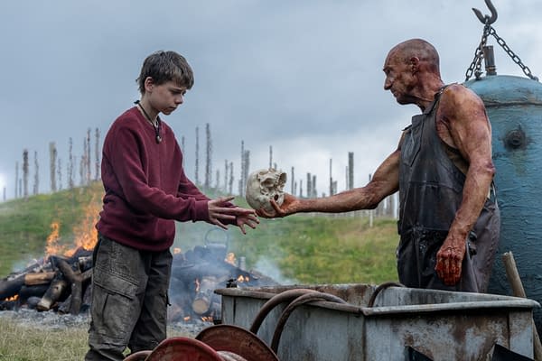 A dramatic scene featuring a young boy in a red sweater and a muscular, blood-stained man exchanging a skull, set against a backdrop of smoky flames and blurred silhouettes of trees. The image conveys a tense interaction amidst a grim environment.