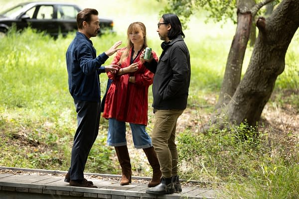 Three people engaged in conversation outdoors, surrounded by greenery; one person wears a red patterned top and brown knee-high boots, while another is in a navy jacket, and the third wears a black jacket. A black car is visible in the background.