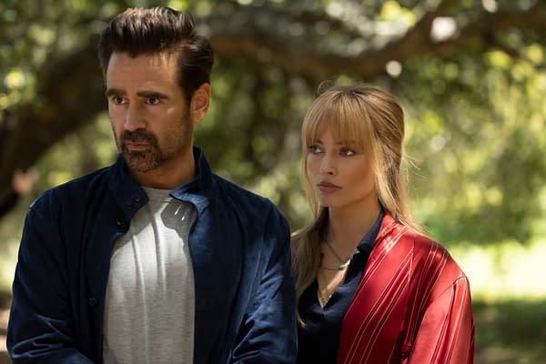 A scene featuring two people standing close together in a sunlit outdoor setting, with the man dressed in a dark blue jacket over a gray shirt, and the woman in a red patterned wrap over a dark top. The background shows soft-focus greenery, creating a serene ambiance.