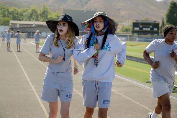 A scene from Disney's FREAKIER FRIDAY showing Julia Butters as Harper Coleman and Sophia Hammons as Lily Davies, both wearing athletic outfits and accessories while jogging on a track, with other runners in the background.