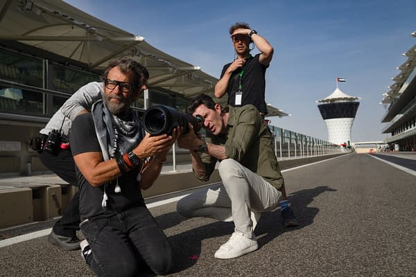 A group of photographers captures the action on a racetrack, with one man in the foreground kneeling and aiming a large camera lens, while another photographer adjusts his focus in the background. The iconic control tower of a Formula 1 race is visible in the distance.