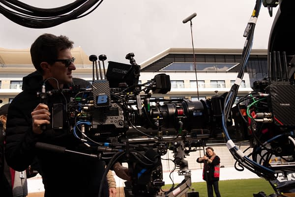 A film crew member operates a professional camera setup on a film set, with various cables and equipment visible, while another crew member prepares in the background. The setting appears to be an outdoor venue, suggesting a dynamic production environment.