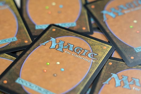 Colorful backs of Magic: The Gathering trading cards are stacked together, featuring an ornate design with a prominent logo and various colored dots, emphasizing the fantasy and gaming theme.