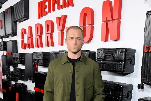 A man stands in front of a backdrop featuring the words "Netflix CARRY ON" in bold red letters, surrounded by black luggage cases. He is wearing an olive green jacket over a black shirt, displaying a serious expression.