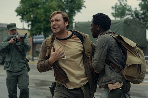 A tense moment from 'The Long Walk,' featuring two characters: one with a distressed expression and a plaid shirt, and another looking concerned, both wearing backpacks. In the background, a soldier is aiming a firearm, adding to the scene's intense atmosphere.