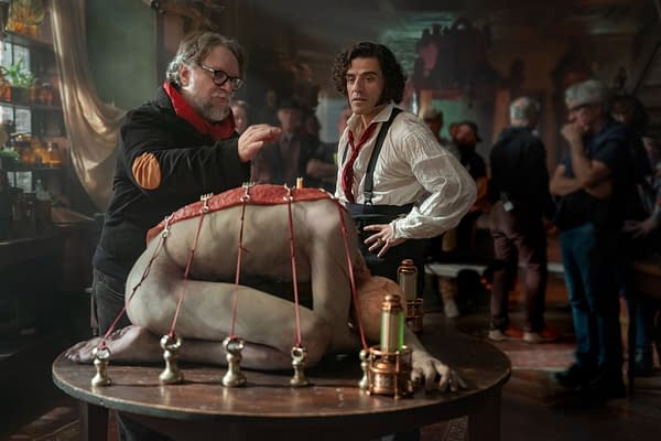Behind-the-scenes image from 'Frankenstein,' featuring director Guillermo del Toro instructing actor Oscar Isaac, who portrays Victor Frankenstein, beside a grotesque creation on a table. The setting is dark and atmospheric, filled with props related to the film.