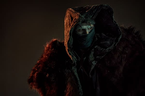 Jacob Elordi portrays The Creature from 'Frankenstein,' wearing a dark hooded cloak and a scarf that covers part of his face, in a dramatic and moody setting.