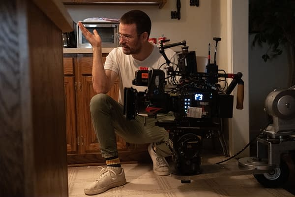 A man with glasses is crouched on the floor in a kitchen, engaged in conversation while gesturing with his hand. Next to him, a professional video camera is set up, indicating a filming scene.