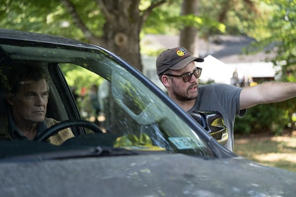 A scene from a film set featuring a serious actor in the driver's seat of a car, while a director gestures animatedly outside, suggesting directions. The background shows trees, indicating an outdoor setting.