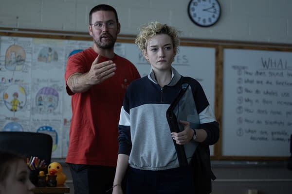 A teacher in a red shirt gestures while speaking to a student with curly hair, who holds a folder. In the background, a classroom is visible with various drawings and a clock on the wall.