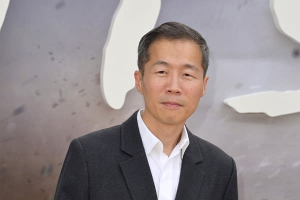 A man with short black hair is wearing a gray blazer over a white shirt, standing against a blurred background. He has a neutral expression and appears poised.