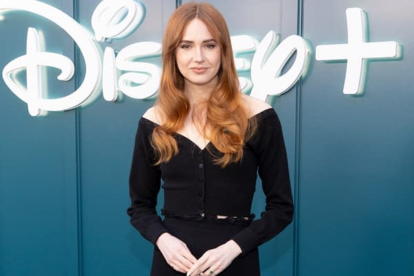 A woman with long, wavy reddish-brown hair stands in front of the Disney+ logo. She is wearing a black off-shoulder top and has a relaxed expression, with her hands resting in front of her.