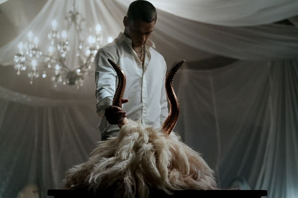 A dramatic scene from the film 'HIM', featuring a character holding a pair of large, twisted horns above a fluffy, fur-like object on a table. The setting is dimly lit, with a chandelier visible in the background, creating a tense and mysterious atmosphere.