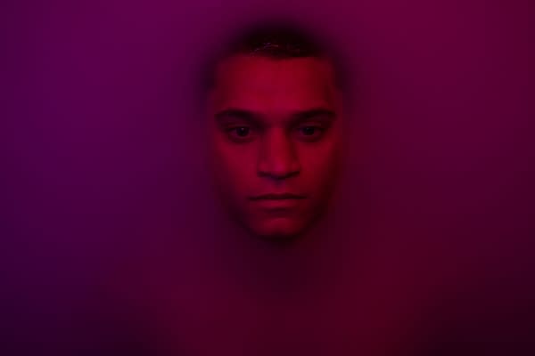 A close-up portrait of a young man, Tyriq Withers, with a serious expression. The image is enveloped in purple tones, creating a dramatic and moody atmosphere, reflecting themes from the film 'HIM', directed by Justin Tipping.