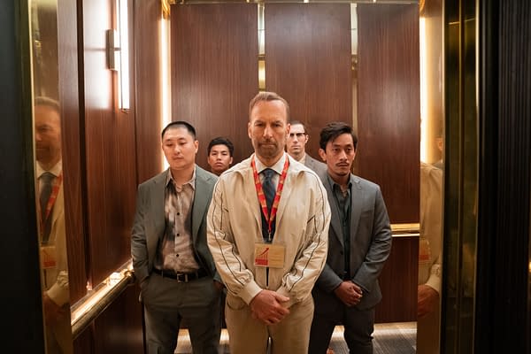 A group of five men stands solemnly in an elevator, dressed in suits and casual attire, with one man prominently wearing a badge around his neck. The setting features polished wooden walls and soft lighting, creating a tense atmosphere.