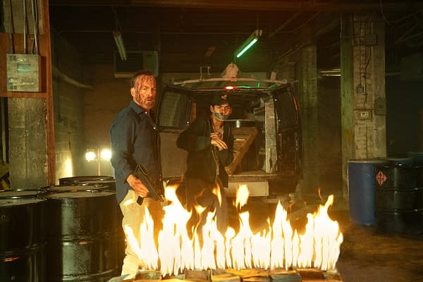 Two men in a dimly lit industrial setting, one holding a gun with a bloodied face, while the other appears to be hiding behind him. Flames erupt in front of them, with black barrels and a vehicle in the background.