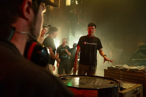 A group of filmmakers collaborates in a dimly lit warehouse setting, with one man gesturing while speaking. The atmosphere is smoky, and various props, including a barrel and stacks of crates, are visible.